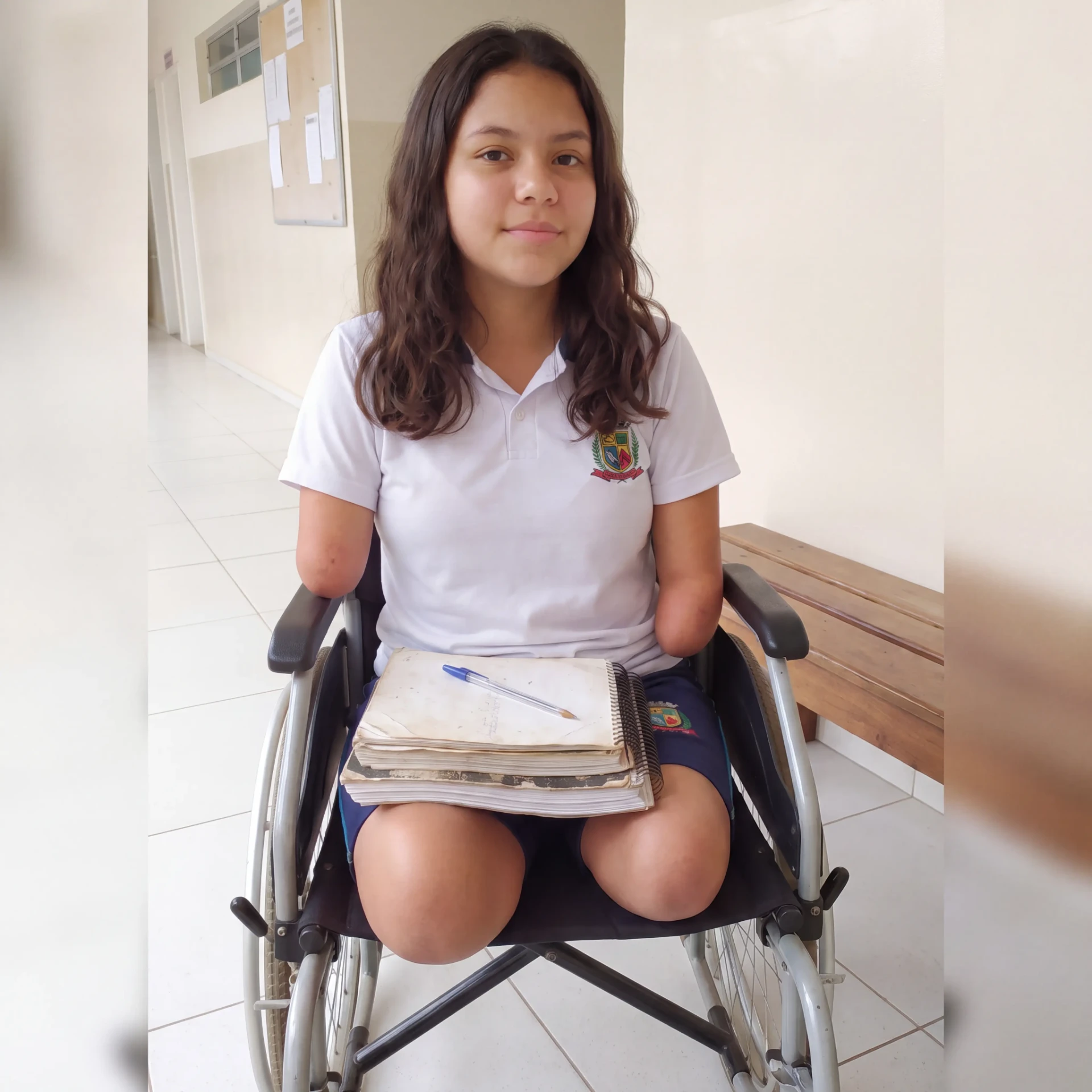 Rebeca sentada na cadeira de rodas da escola, segurando seus cadernos